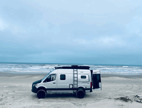 White Winnebago Revel RV rental parked on a sandy beach by the ocean. A dog sits inside the driver's seat, enjoying the view.