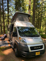 This silver camper van rv rental with a pop-top roof is set up at a sunny forest campsite, featuring an outdoor chair and mat.