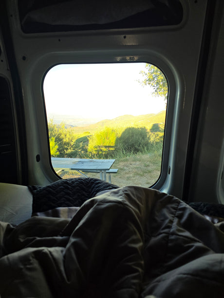 Scenic RV rental morning view from a comfy camper van bed, overlooking sunlit green hills & a picnic table.