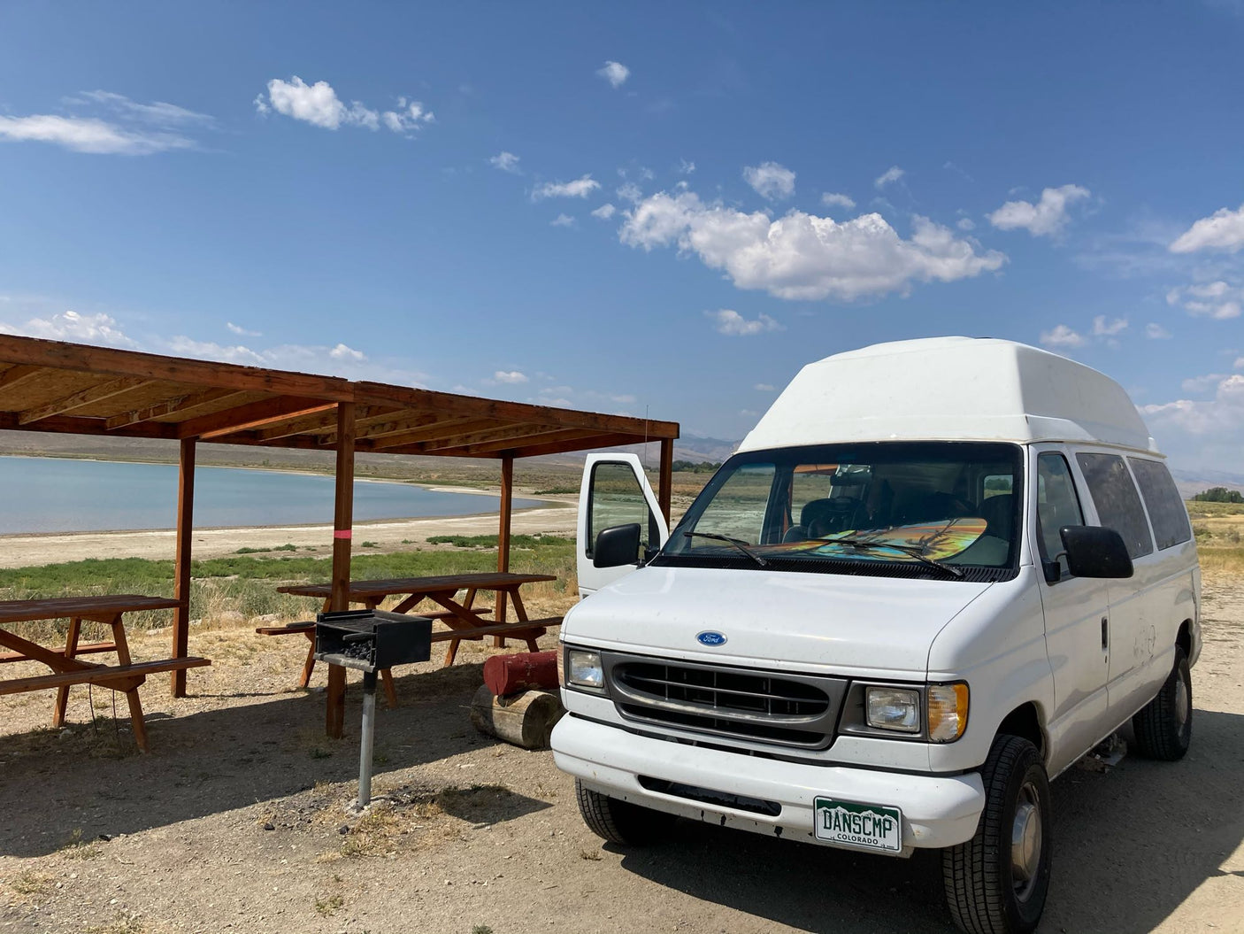 White high-top RV rental van at a scenic campsite with picnic tables, grill, & lake views under blue sky.