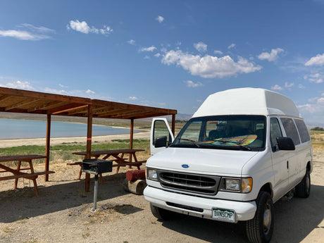 White high-top RV rental van at a scenic campsite with picnic tables, grill, & lake views under blue sky.