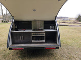 An open RV rental rear hatch reveals a compact outdoor kitchen: sink, stove, microwave, cabinets, and storage. Ideal for camping.