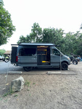 A customized Sandy Vans RV rental parked in nature. This dark grey camper van features an extended awning, bike rack, and lit interior.
