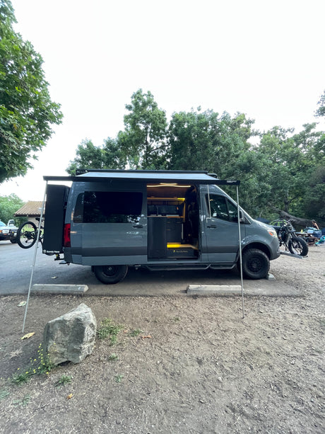 A customized Sandy Vans RV rental parked in nature. This dark grey camper van features an extended awning, bike rack, and lit interior.