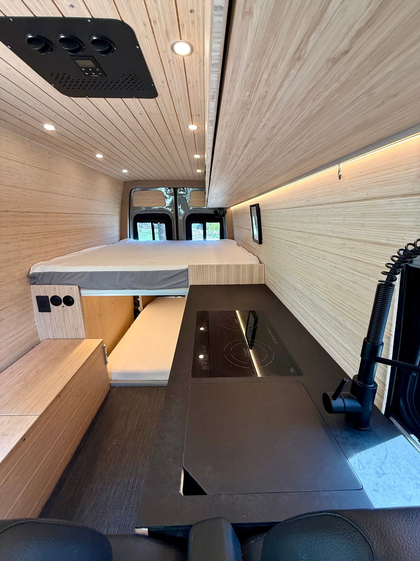 RV rental: Inside a modern camper van featuring a spacious bed, sleek kitchen with cooktop, and warm wood paneling. — photo 8