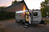 This E-Pro travel trailer (rv rental) is illuminated at dusk in a campsite, with its awning extended and door open, ready for camping.