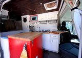 Custom rv rental interior: compact kitchen with light blue microwave, wooden ceiling, bed, and front seats.