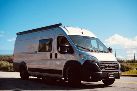 A silver Ram ProMaster camper van, an ideal RV rental, is seen on a road against a clear blue sky.