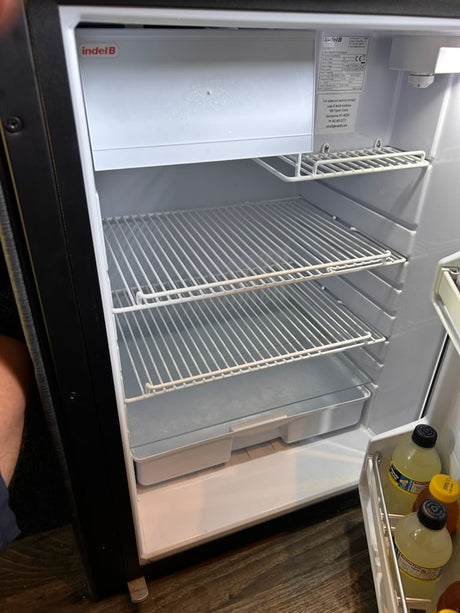 Inside an RV rental's clean, empty compact refrigerator with wire shelves and bottled drinks in the door.
