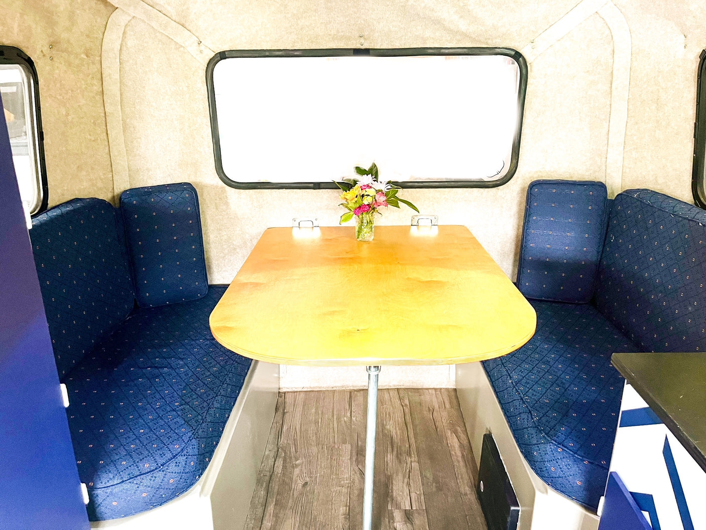 Cozy RV rental interior features blue dinette seating, a wooden table with flowers, and a large window. Ideal for travel.