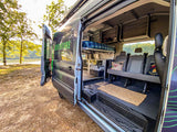 Spacious RV rental camper van with side door open, revealing bunk beds, seating, and fridge by a scenic lake.