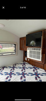 Cozy RV rental interior with a patterned quilt on the bed, wall-mounted TV, AC unit, and wooden cabinets.