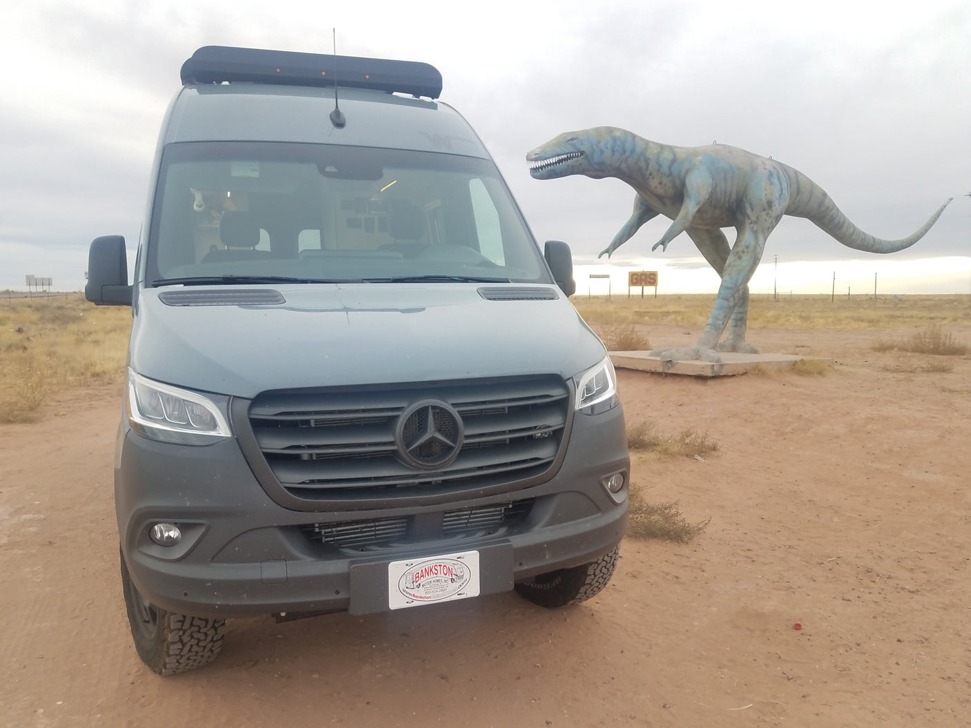 Mercedes-Benz Sprinter RV rental parked near a large dinosaur statue and 'GAS' sign in an arid landscape.