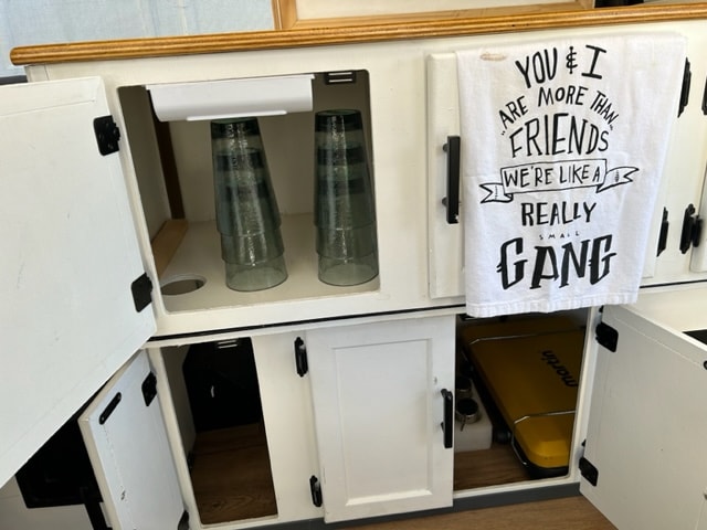 RV rental: Open compact kitchen area, with stacked glasses, hidden storage, and a yellow 'Martin' case. Towel reads 'SMALL GANG'. — photo 9