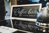 Charming RV rental coffee bar setup: a black 'Coffee bar - OPEN DAILY -' sign with reflection, alongside a kettle and appliance.