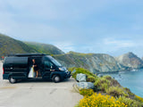 Embrace van life with an RV rental! Woman exits her black camper van on a scenic coastal overlook, mountains in view.