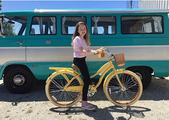 Young girl on a yellow bicycle smiles in front of a teal and beige vintage RV rental van, ready for adventure.