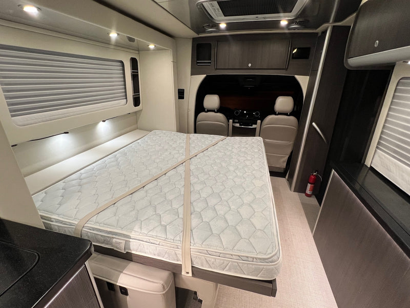 This RV rental interior features a comfy bed with security straps, a fire extinguisher, overhead storage, and front seats for travel. — photo 5