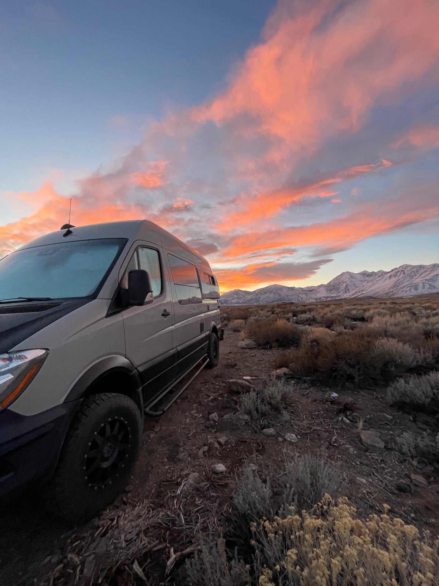 Adventure awaits in this RV rental camper van, parked amidst desert shrubs with a vibrant sunset over snowy mountains.