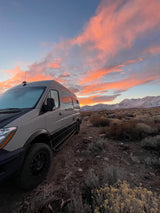 Adventure awaits in this RV rental camper van, parked amidst desert shrubs with a vibrant sunset over snowy mountains.