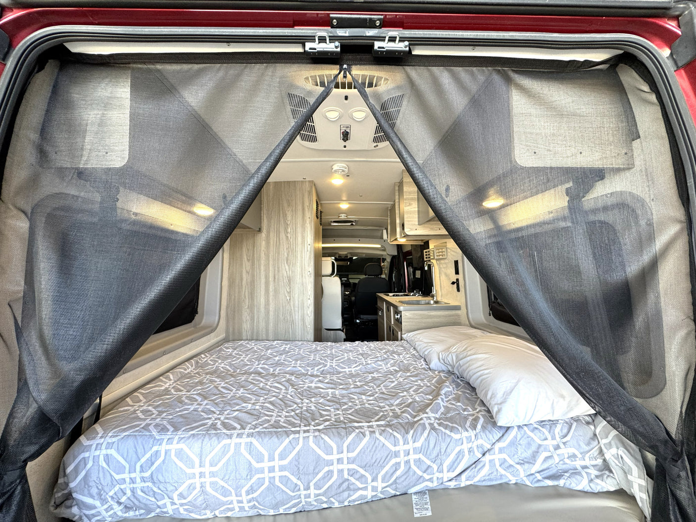 Inside an rv rental: a comfy bed with a geometric comforter, a bug screen at the rear, and a glimpse of the kitchen.