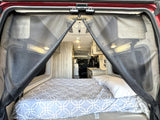 Inside an rv rental: a comfy bed with a geometric comforter, a bug screen at the rear, and a glimpse of the kitchen.