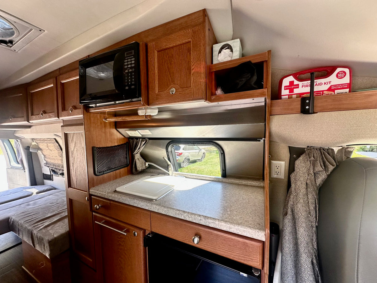 Compact rv rental interior: kitchenette with microwave & sink, overhead cabinets, and a first aid kit. Sleeping area visible. — photo 5