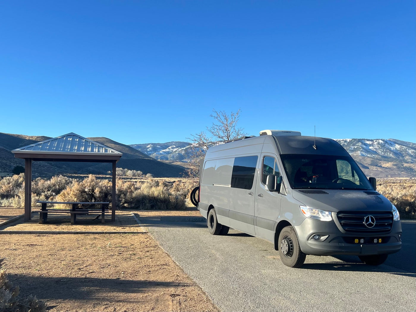 Explore nature in this gray Mercedes Sprinter rv rental, parked by a picnic shelter with snowy mountains in the background. — photo 6