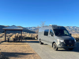 Explore nature in this gray Mercedes Sprinter rv rental, parked by a picnic shelter with snowy mountains in the background.