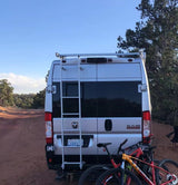 Rear view of a silver RV rental camper van with a ladder & two mountain bikes on a bike rack, parked on a dirt road amidst nature.