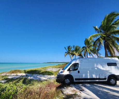 Perfect RV rental getaway: A white camper van parked on a sandy beach next to clear turquoise water and palm trees.