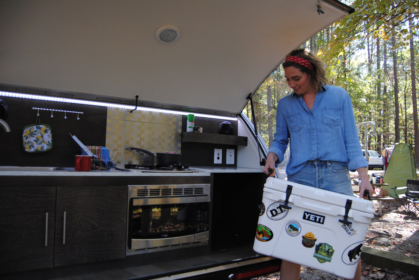 This RV rental features an outdoor kitchen. A woman loads a cooler adorned with National Park stickers, ready for campsite cooking.