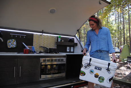 This RV rental features an outdoor kitchen. A woman loads a cooler adorned with National Park stickers, ready for campsite cooking.