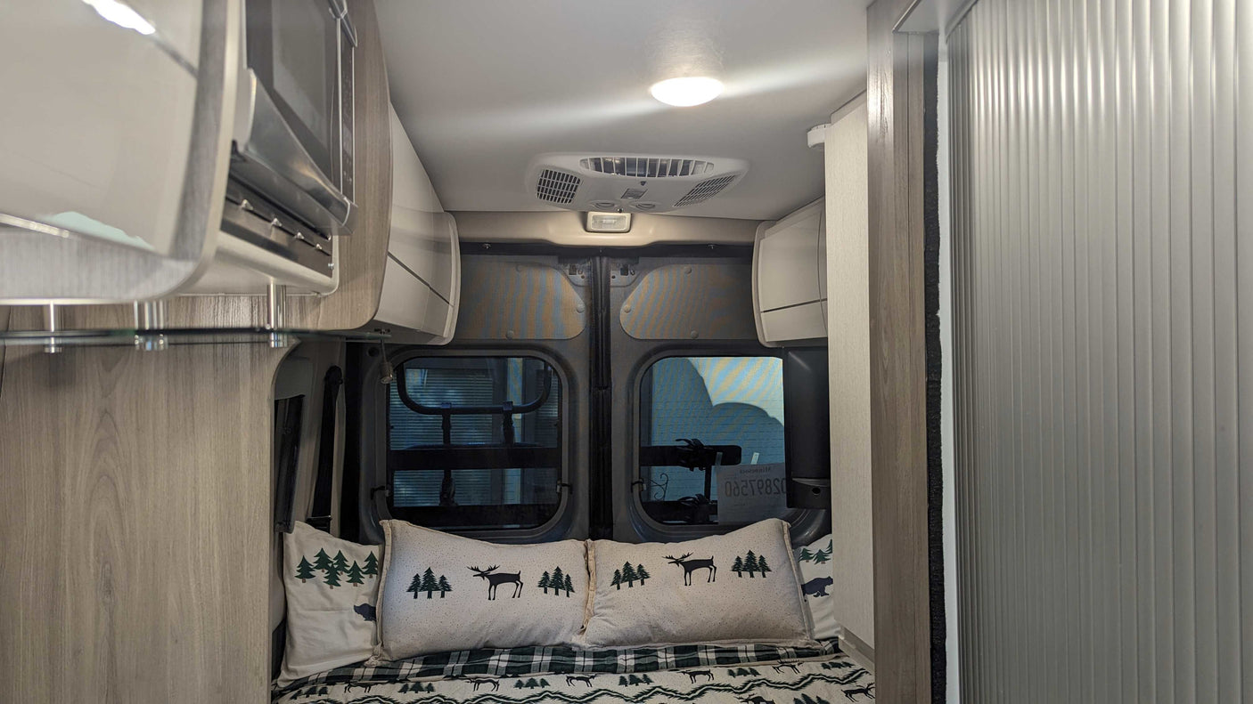 Interior of an RV rental showing a neatly made bed with forest-themed pillows, rear windows, overhead storage, and an AC unit.
