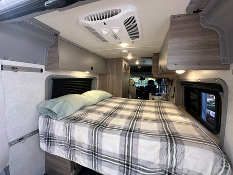 View of a modern RV rental interior featuring a bed with plaid bedding, overhead storage, and ceiling AC, extending to the kitchen.