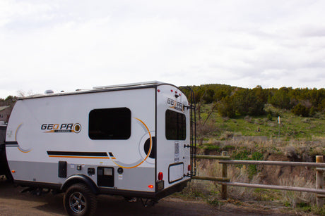 A white and grey GEO PRO travel trailer, available for rv rental, parked outdoors near a fence and trees.