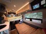 Cozy RV rental interior: an elevated bed, lower sleeping area, kitchenette, and control screen, all with a serene forest view.