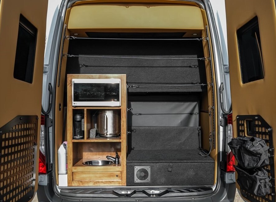 Rear view of a custom RV rental, showcasing a compact mobile kitchen with microwave/sink, ample storage, and bed platform for van life. — photo 17