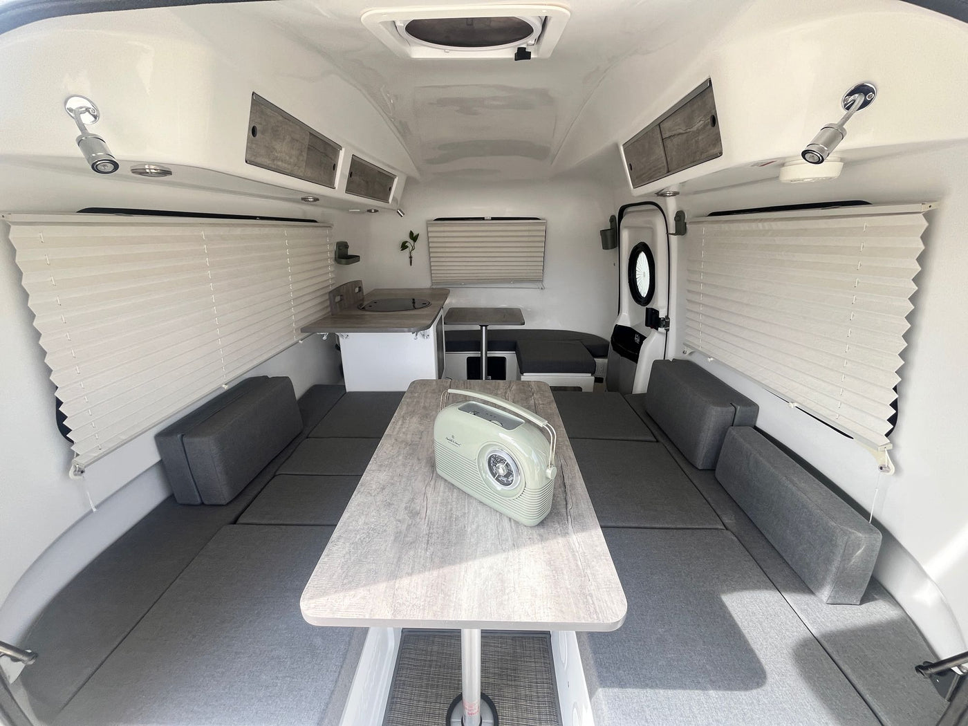 A clean, bright rv rental interior featuring flexible gray cushion seating, a central table, and a small kitchen. — photo 16