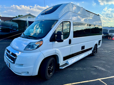 A modern white National Traveler RV rental, with a raised roof and black wheels, is parked on an asphalt lot under a cloudy sky.