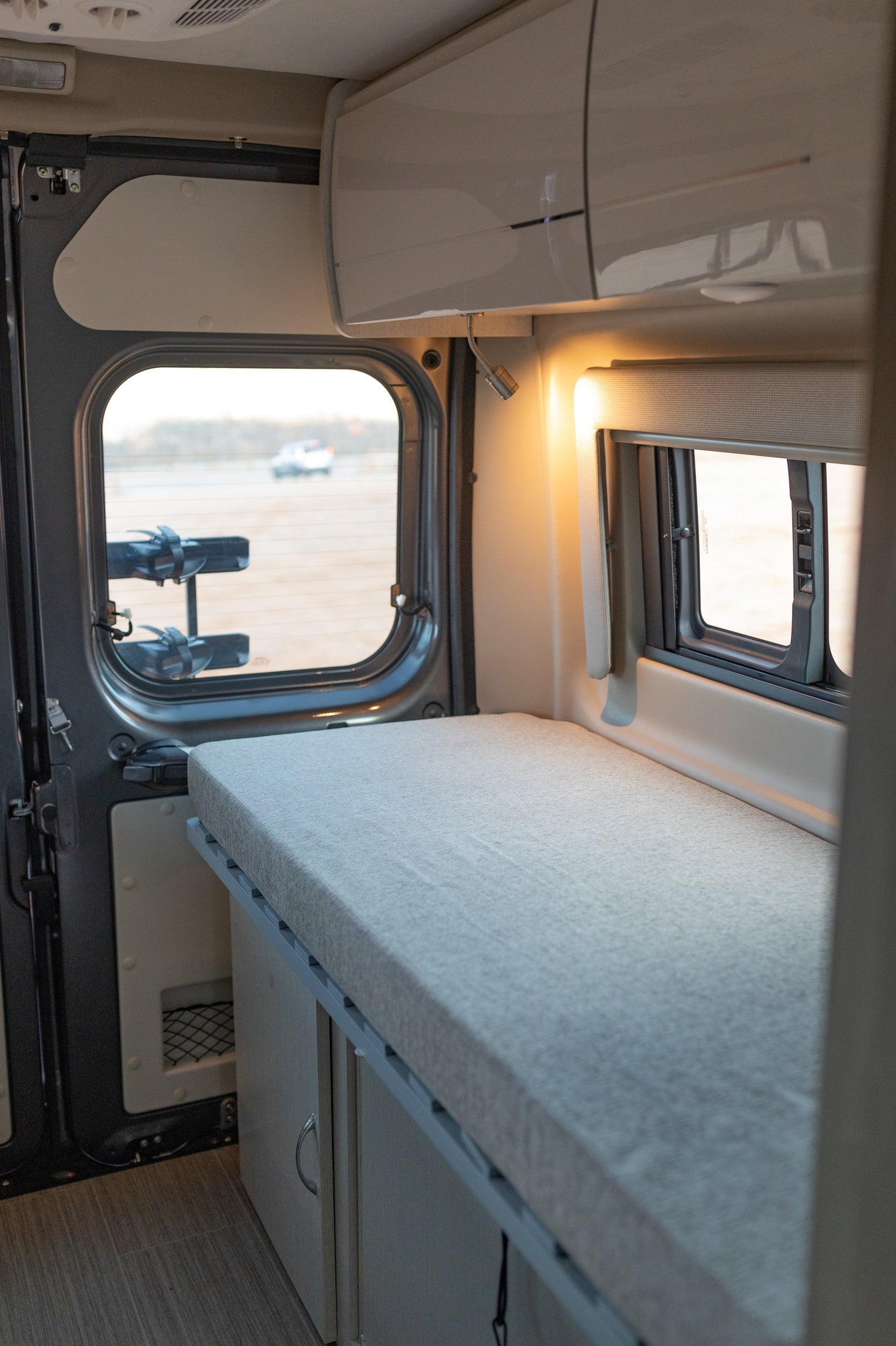 Cozy RV rental interior: light gray bed, clever storage, and windows revealing outdoor views. Perfect for your next adventure.