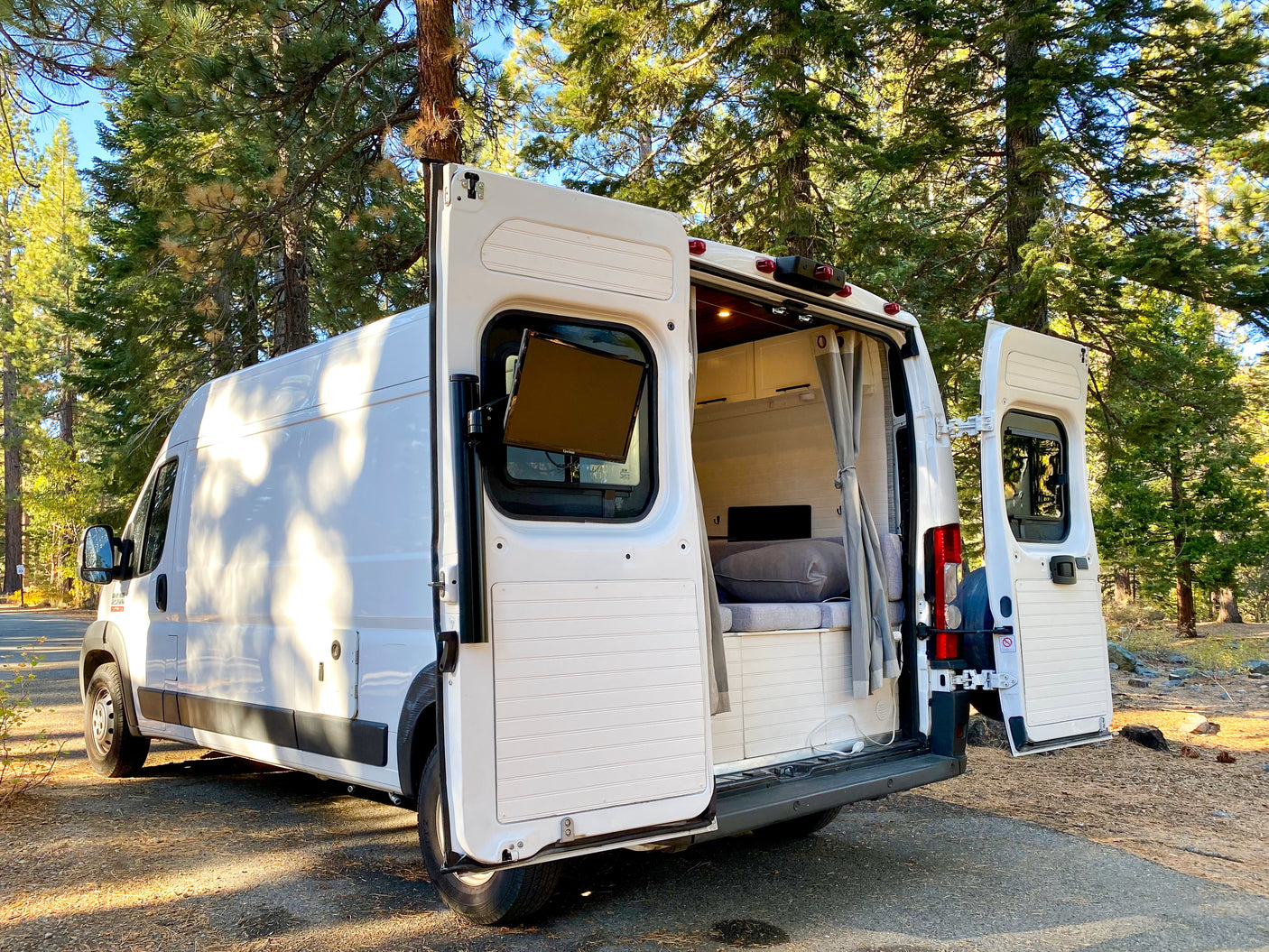 White converted camper van, a great RV rental option, parked in a forest with open doors revealing its cozy interior.
