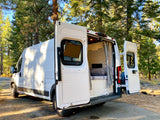 White converted camper van, a great RV rental option, parked in a forest with open doors revealing its cozy interior.