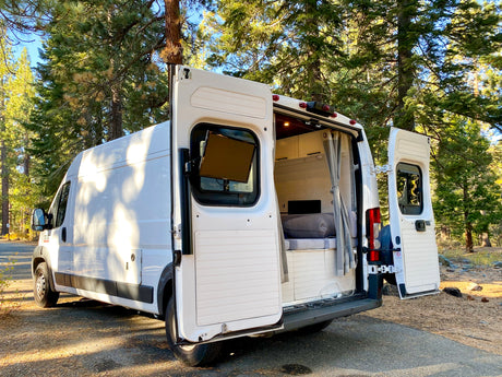 White converted camper van, a great RV rental option, parked in a forest with open doors revealing its cozy interior.