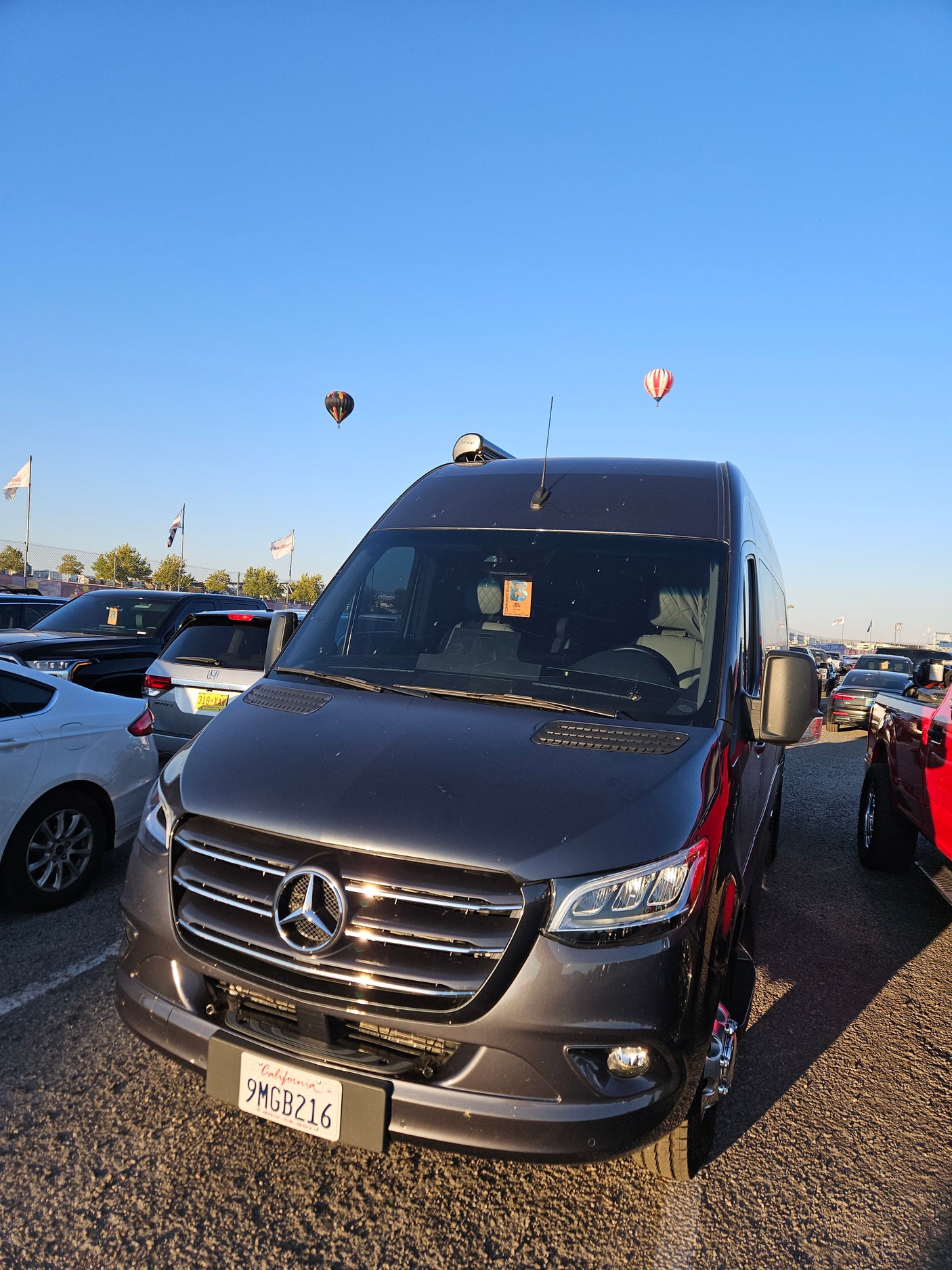 Front view of a dark grey Mercedes-Benz Sprinter RV rental van parked. Two hot air balloons float in the blue sky.