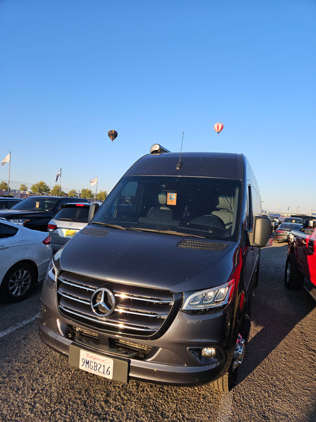Front view of a dark grey Mercedes-Benz Sprinter RV rental van parked. Two hot air balloons float in the blue sky.