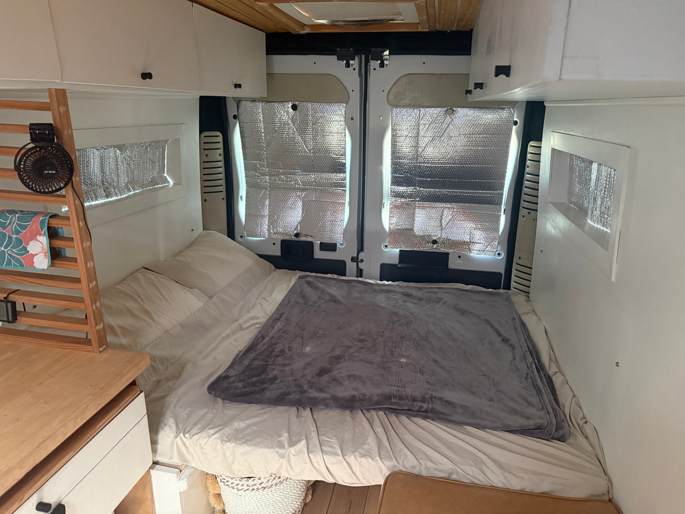 Inside an RV rental van: a bed with a gray blanket, overhead cabinets, and insulated rear windows.