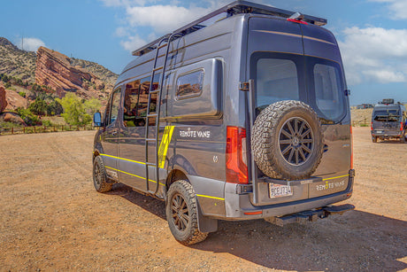 Gray 'REMOTE VANS' adventure RV rental camper van, with off-road tires and roof rack, parked at Red Rocks.