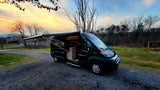Black camper van for rv rental, parked on gravel with open side door & extended awning, set against a vibrant sunset.