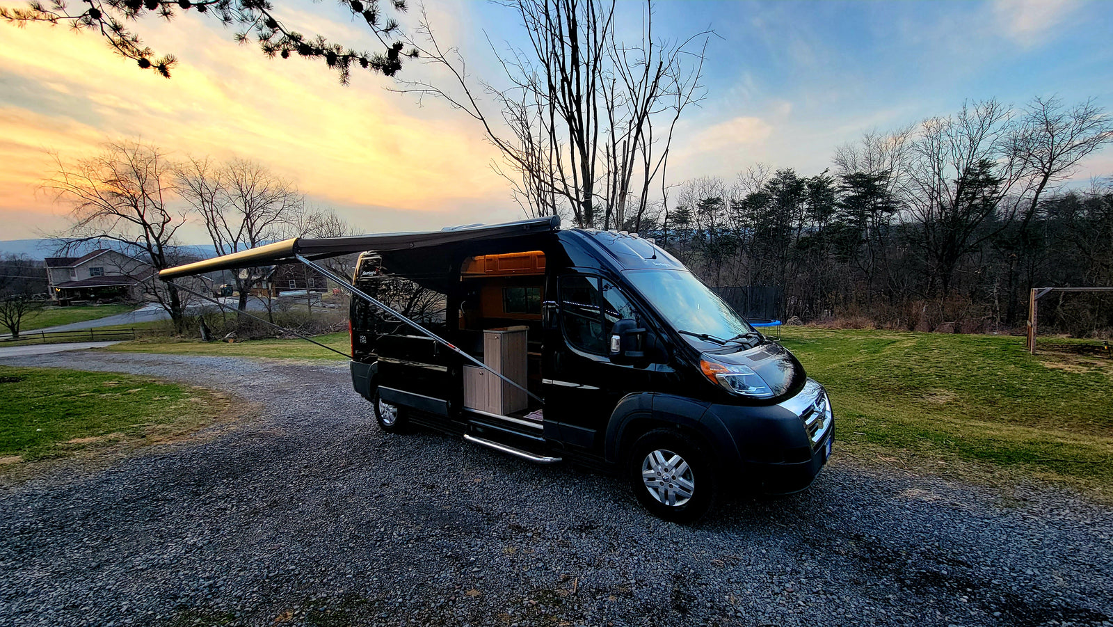 Black camper van for rv rental, parked on gravel with open side door & extended awning, set against a vibrant sunset.